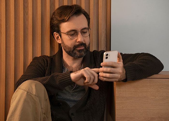 Man with glasses and beard sitting indoors, looking at his smartphone, reflecting privacy concerns and trust issues.