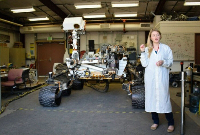 A woman in a lab coat standing next to a large Mars rover model showing human for scale indoors.