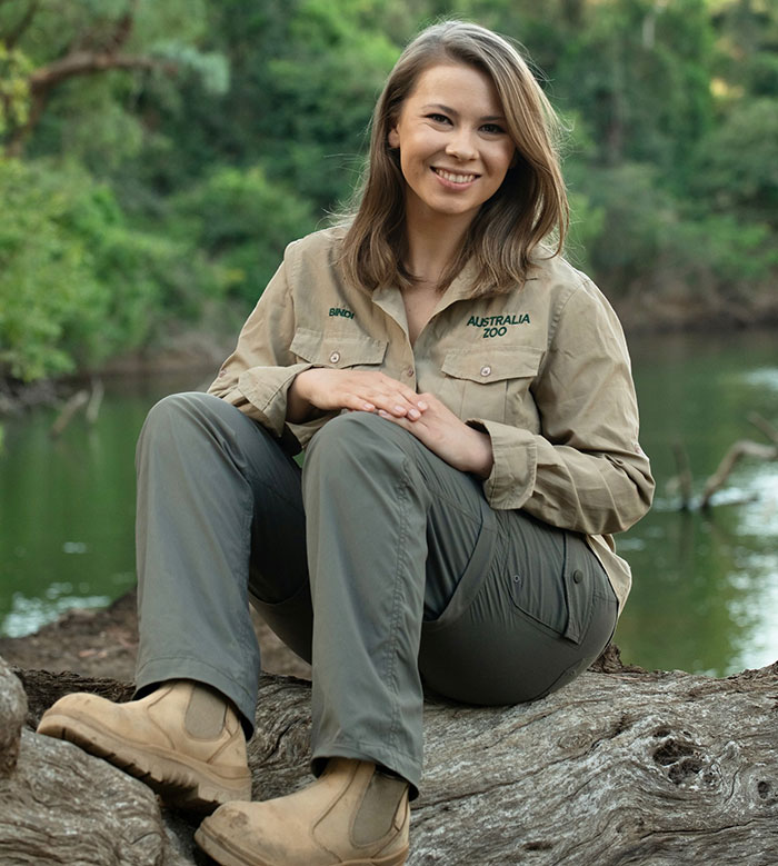 Bindi Irwin sitting outdoors by a river, smiling and wearing Australia Zoo uniform, announcing move from Australia. Bindi Irwin sitting outdoors by a river, smiling and wearing Australia Zoo uniform, announcing move from Australia.