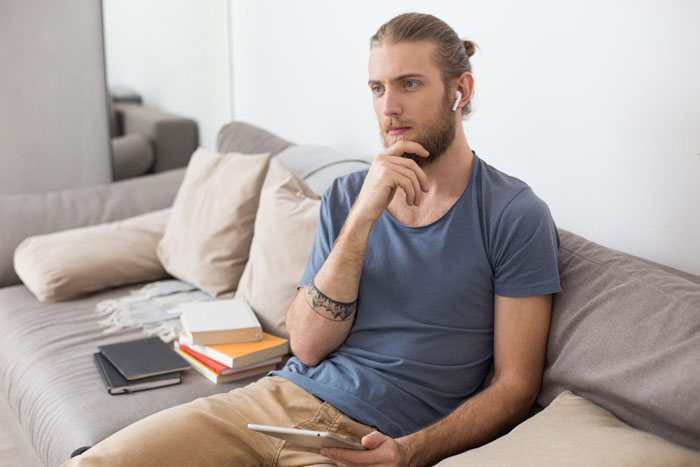 Man with tattoo sitting on couch holding tablet, looking thoughtful after birth family tracks him down