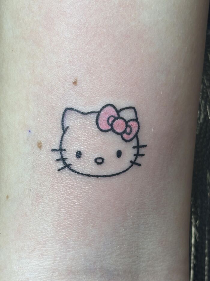 Simple Hello Kitty tattoo in black outline with pink bow on light skin, showcasing tattoo artist work with challenging client requests.