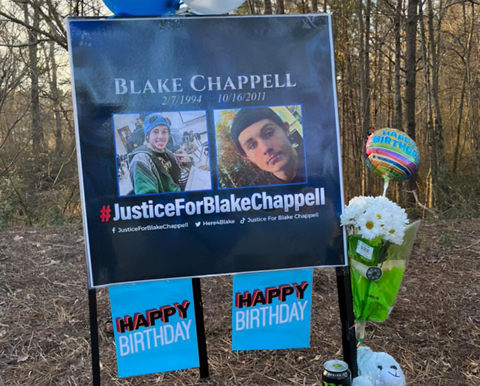 Memorial sign and flowers honoring Blake Chappell with JusticeForBlakeChappell hashtag in cold case breakthrough setting.