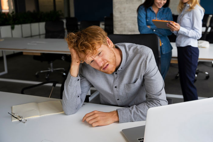 Frustrated man at office desk with laptop looking stressed while two women in background discuss boss invite vacation with her.