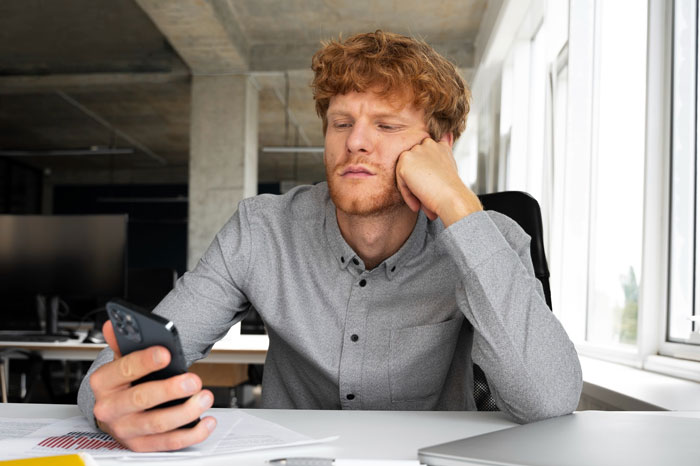 Young man in a gray shirt looking frustrated at his phone, possibly dealing with a boss pretending never gave notice situation.