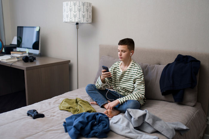 Teen boy with earphones sitting on bed, using smartphone, surrounded by scattered clothes in a casual bedroom setting.
