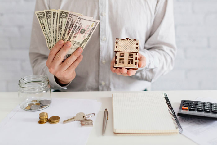 Man holding US dollar bills and a small wooden house model symbolizing son&rsquo;s survivor benefits and financial decisions.