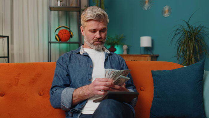 Middle-aged man counting money on an orange couch, illustrating themes of survivor benefits and relationship conflict.