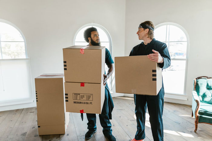Two men carrying moving boxes in a bright room illustrating a woman dumps BF after being told her period is disgusting.