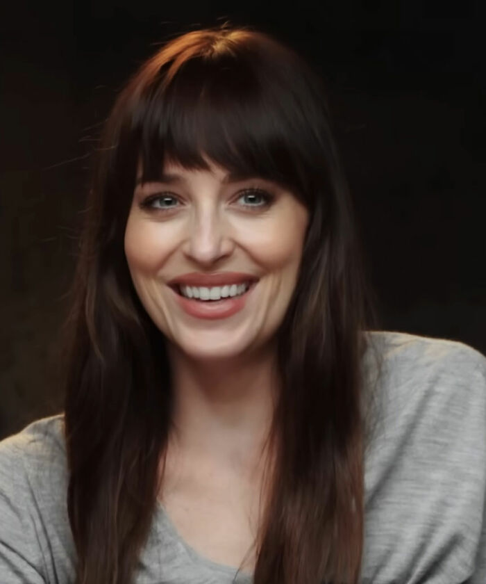 Dakota Johnson smiling with long brown hair and bangs, wearing a casual gray top in a dimly lit setting.
