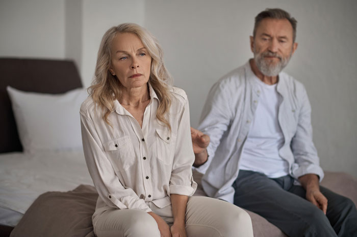 Older couple sitting on bed looking upset, woman avoiding man reaching out, symbolizing breaking up over photograph.