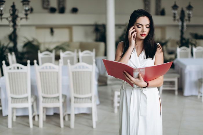 Bride in white dress on phone, reviewing wedding seating chart in a decorated room with white chairs and tables.