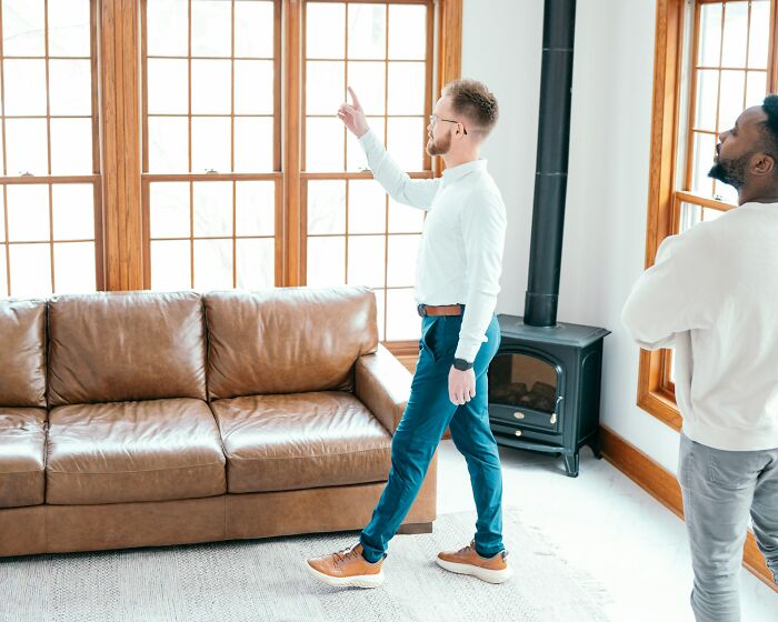 Two men in a living room discussing out of touch things people have been told near a brown leather couch and windows.