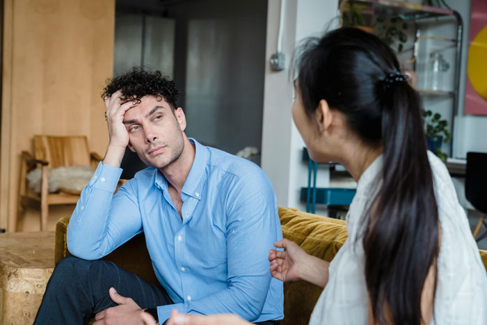 Man in blue shirt sitting on couch looking tired while woman with long black hair talks to him indoors