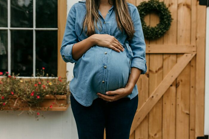 Pregnant woman in casual blue shirt standing outdoors near wooden door and window with flowers.