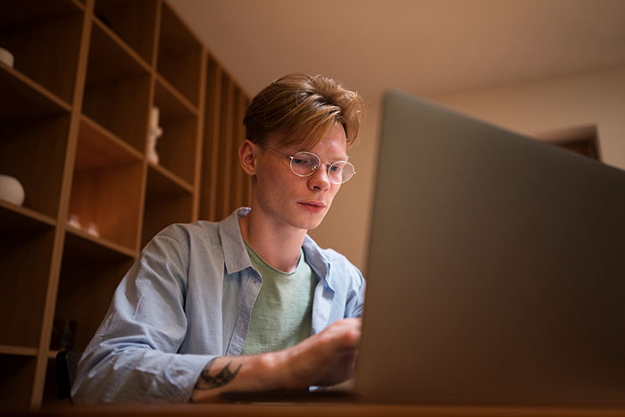 Young man wearing glasses, focused on his laptop, possibly reading or responding to catfish stories in a quiet room.