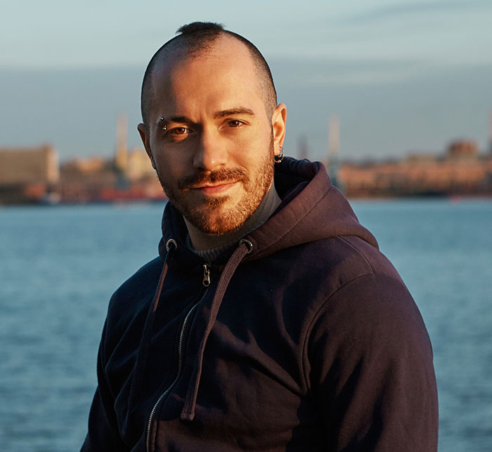Man with shaved head and beard wearing a hoodie near water, evoking feelings connected to catfish stories and online trust issues.