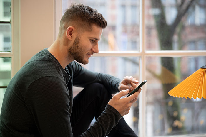 Young man sitting by a window, focused on his phone, reflecting on catfish stories and online DM experiences.
