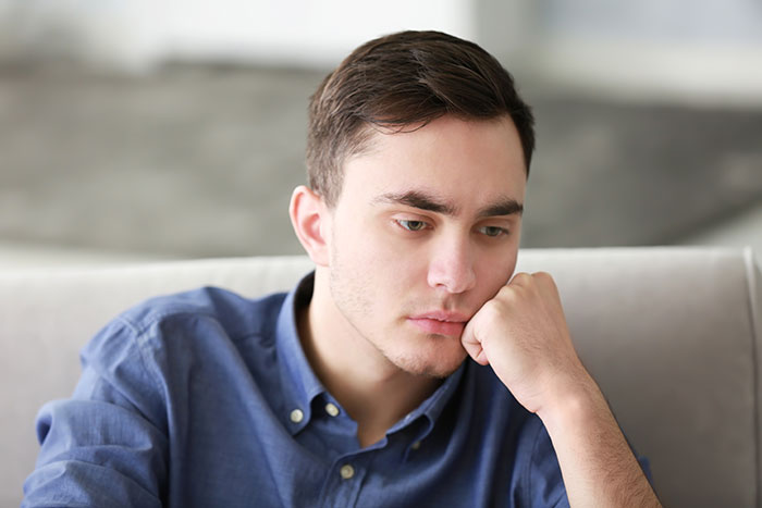 Young man looking thoughtful and guilty while sitting on a couch, relating to catfish stories and online deception.