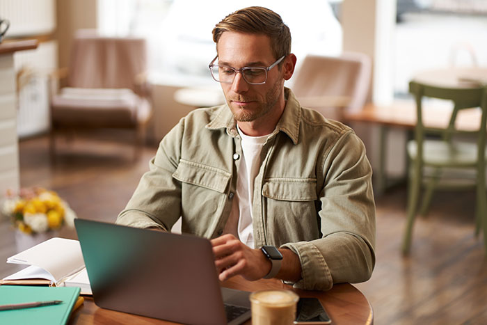Man wearing glasses and a jacket using laptop at table, reflecting on catfish stories and online DMs suspicion.