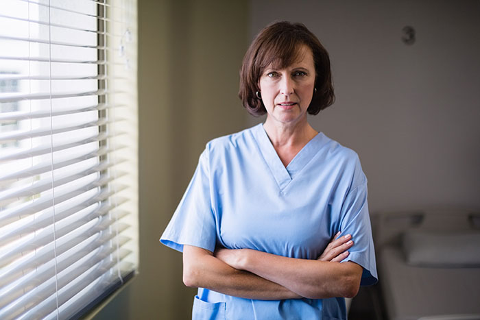 Woman in blue medical scrubs standing with arms crossed near window, reflecting on catfish stories and online messages.