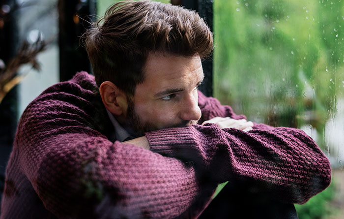 A man in a purple sweater looking out a rainy window, reflecting on catfish stories and online trust issues.