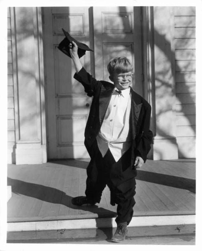 Young child star in oversized graduation gown and cap, smiling and celebrating outside a building, representing child stars.
