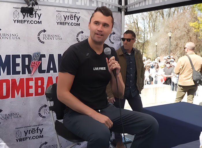 Charlie Kirk speaking at an event, holding a microphone, with a banner showing Turning Point USA and American comeback logos.