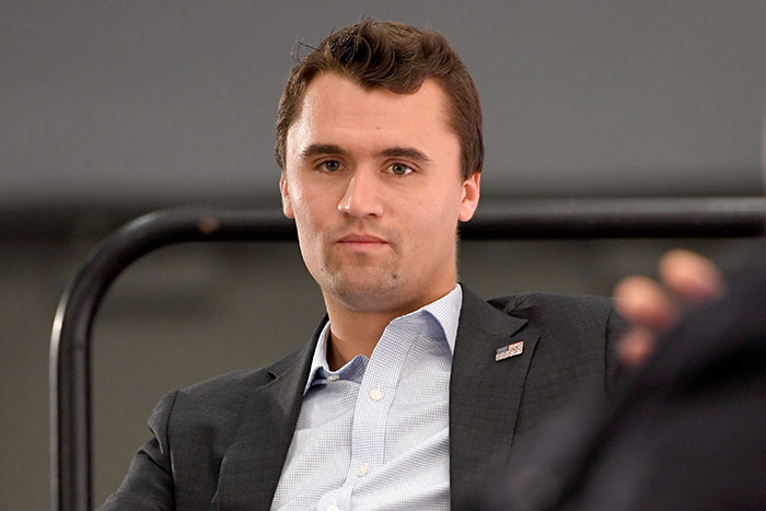 Charlie Kirk wearing a dark suit and light shirt, seated indoors, with a serious expression during an event.