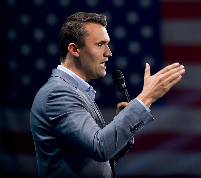 Man in a gray suit holding a microphone speaking passionately with an American flag backdrop in a public event setting.