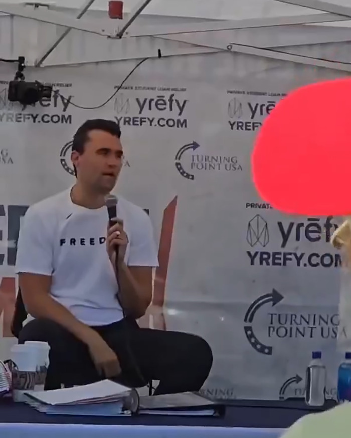 Charlie Kirk speaking at a Turning Point USA event, holding a microphone and seated in front of a branded backdrop.