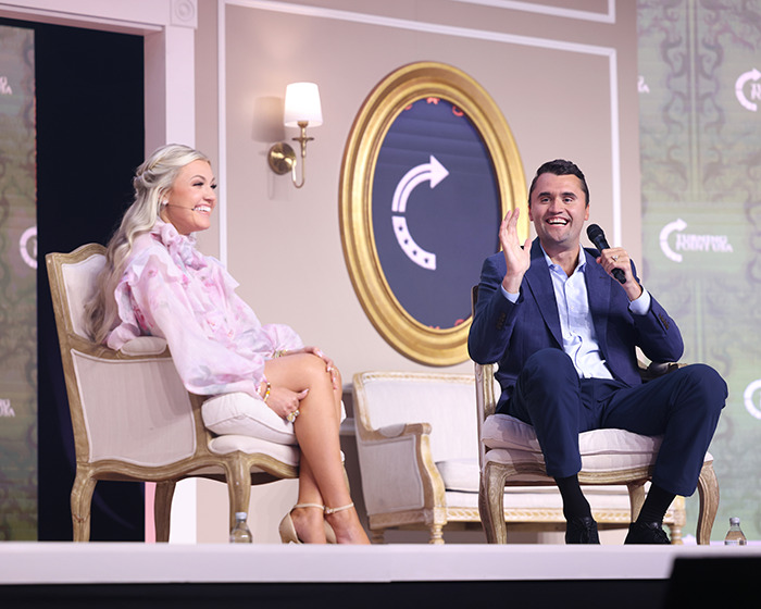Erika Kirk seated on stage in a pink blouse during a Turning Point USA event with a male speaker holding a microphone