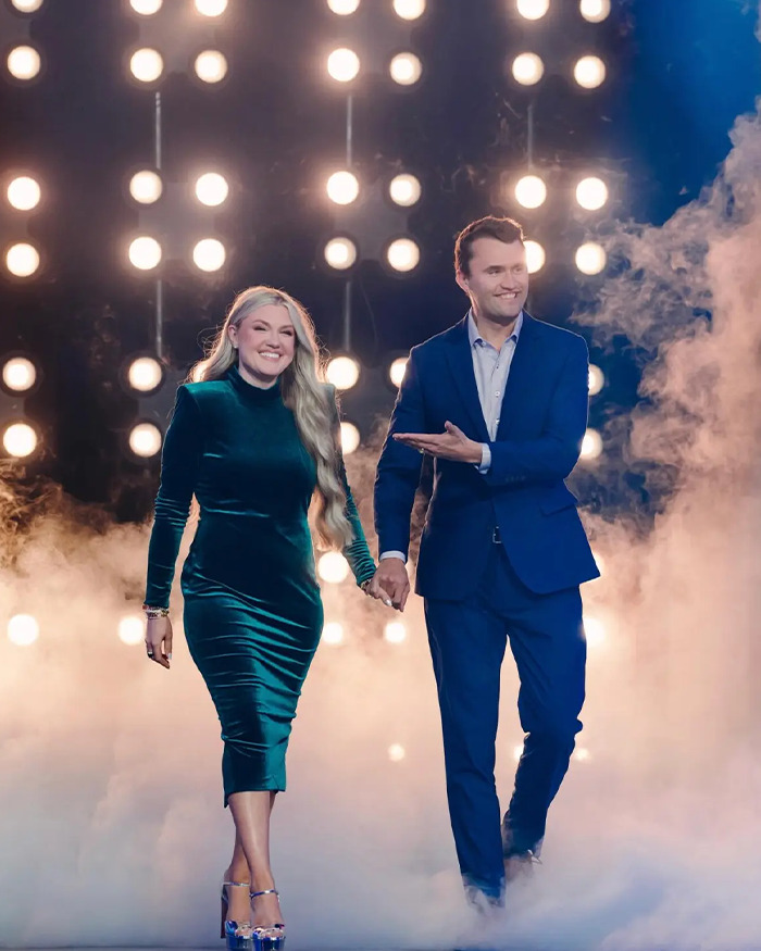 Erika Kirk and a man walking hand in hand on a foggy stage with bright lights, highlighting Turning Point USA CEO.