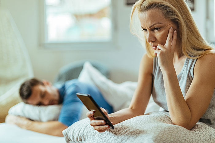 Woman looking concerned at phone while man sleeps in background, depicting husband finds out wife is cheating and revenge.