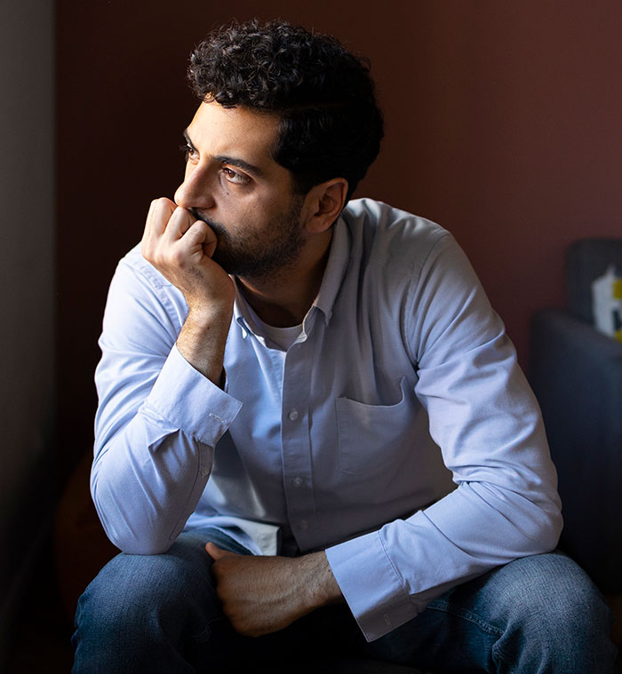 Man in a blue shirt sitting thoughtfully by a window, symbolizing a husband dealing with cheating and planning revenge.