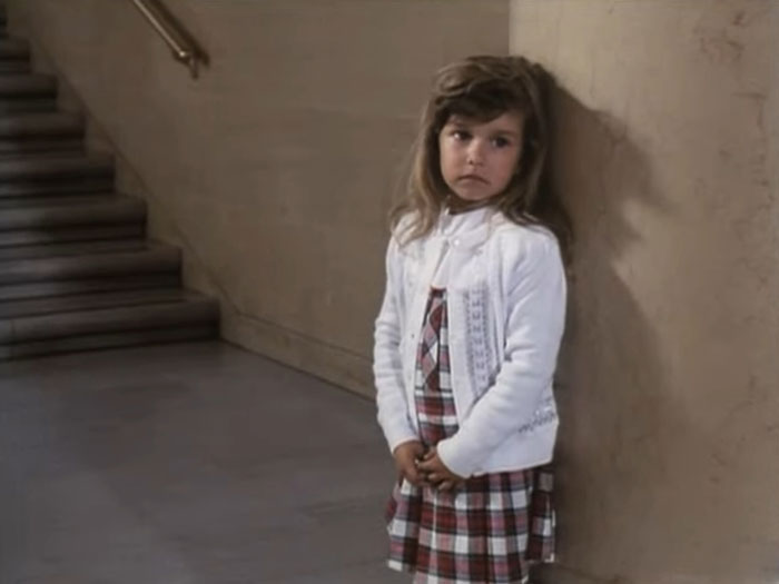 Young child star standing alone near stairs, wearing a plaid dress and white cardigan, reflecting tragic child stars history.