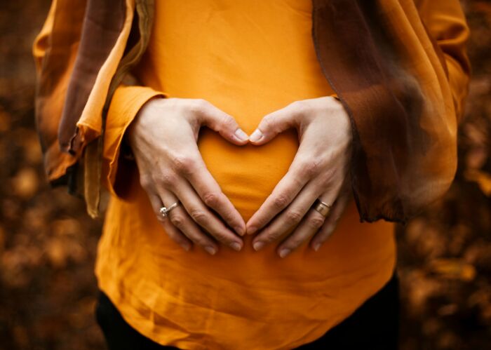 Pregnant mother wearing orange making heart shape with hands over belly, symbolizing child and motherhood connection. Pregnant mother wearing orange making heart shape with hands over belly, symbolizing child and motherhood connection.