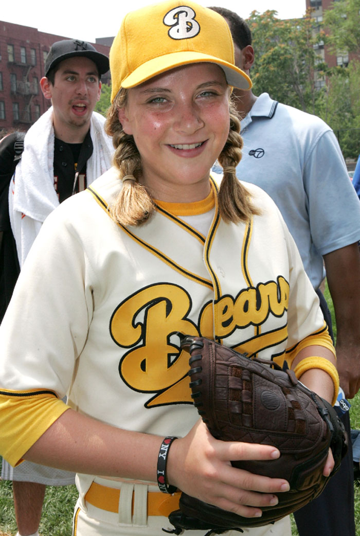 Young child star in a Bears baseball uniform smiling and holding a glove during an outdoor event with fans behind her.