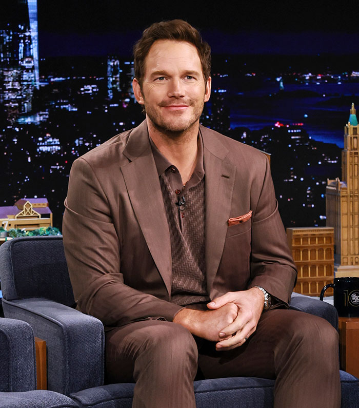 Chris Pratt sitting on a talk show set wearing a brown suit, smiling during an interview at night.