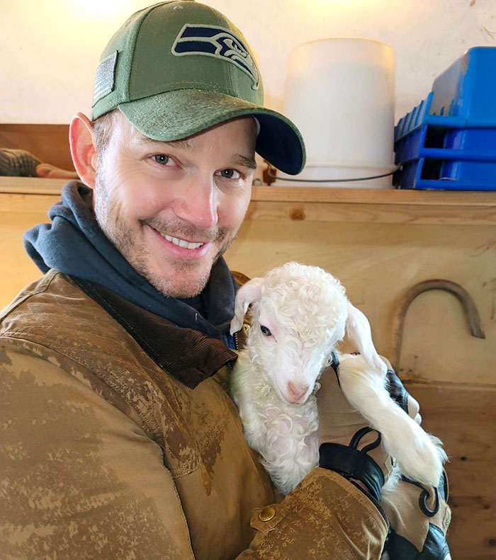 Chris Pratt smiling while holding a small white lamb indoors, related to viral clip of his unusual prayer style.