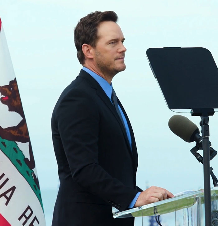 Chris Pratt speaking at a podium with a microphone and California flag, criticized for his unusual way of praying.