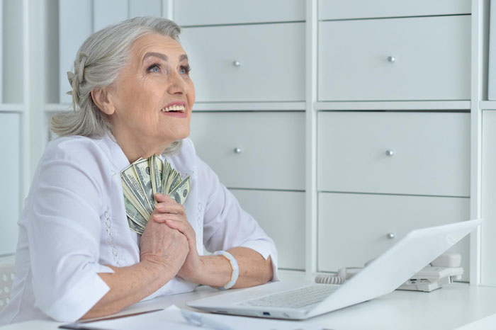 Elderly woman holding dollars with a hopeful expression, sitting at a desk near a laptop, symbolizing Christian rapture.