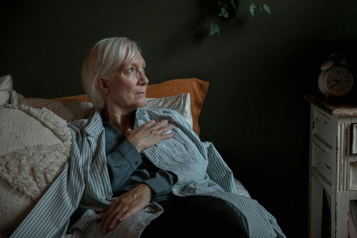 Older woman in striped pajamas sitting on bed with hand on chest, reflecting on Christian rapture and money concerns.