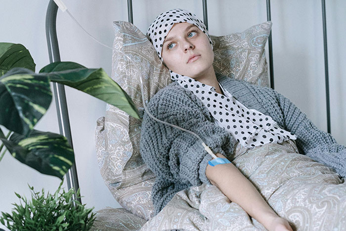 Young chronically ill woman resting in bed with IV drip, wearing a headscarf and cozy gray sweater, looking thoughtful.