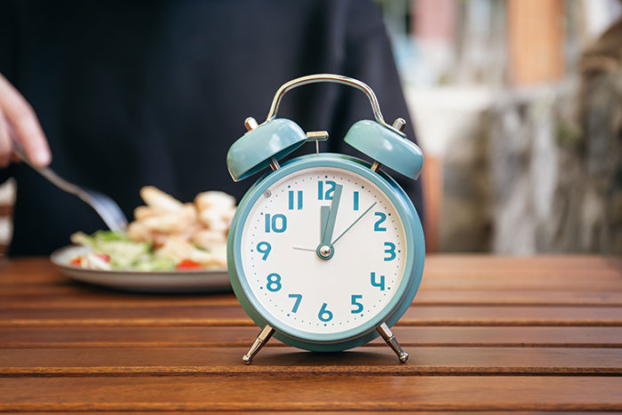 Blue alarm clock on a wooden table with a blurred person eating, representing ridiculous cures for chronically ill people.