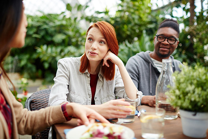 Three friends discussing ridiculous cures for chronic illness while sitting at a wooden table outdoors with drinks and plants.