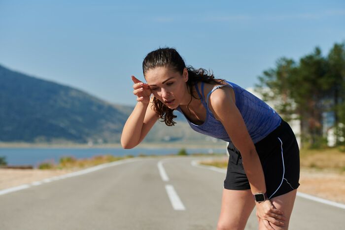 Woman in workout clothes bent over on road, appearing out of breath after exercise near mountains and water, dumbest things concept.