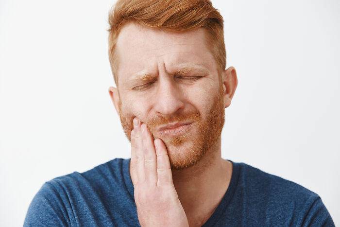 Red-haired man with a beard, wearing a blue shirt, holding his cheek in pain, depicting real-life hexes and curses.