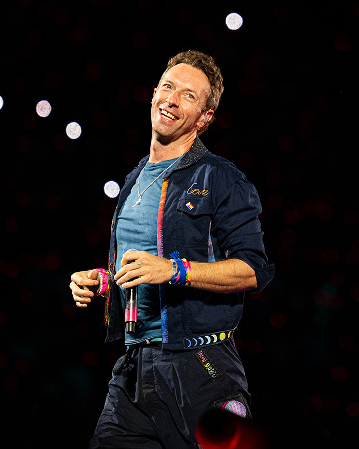 Chris Martin performing on stage, wearing colorful bracelets and a navy jacket during a live concert moment.