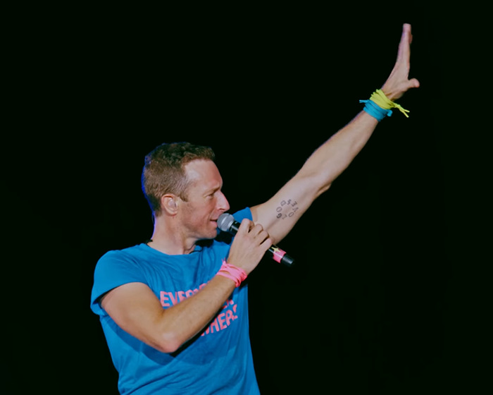 Coldplay&rsquo;s Chris Martin speaking into a microphone on stage, raising his arm during a live concert moment.