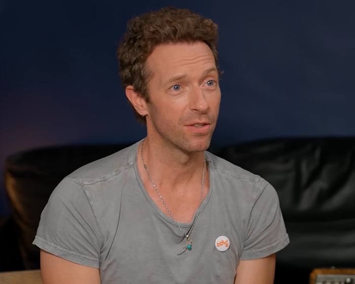 Chris Martin of Coldplay speaking during an interview, wearing a gray shirt with necklaces in a casual setting.